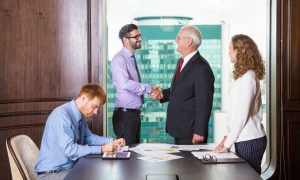 Smiling senior and young businessmen standing at head of table and shaking hands. Businesswoman is standing at table and looking at them. Another businessman is sitting at table and making notes.